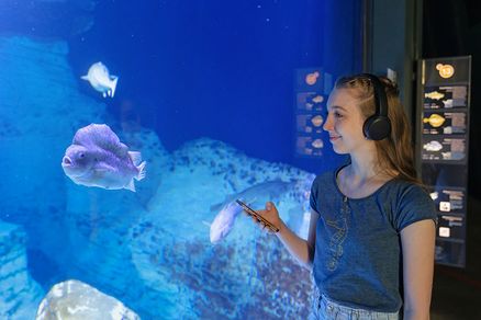 Eine junge Frau mit Kopfhörern und Mobiltelefon steht vor einem Aquarium, in dem Fische schwimmen.