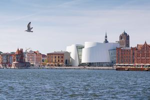 Blick über das Meer auf eine Stadtsilhouette. An der Hafenkante stehen mehrere Backsteingebäude, dazwischen ein modernes weißes Gebäude