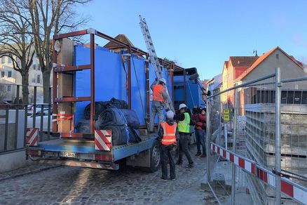 Blick auf einem LKW mit in blauer Folie eingepackten Aquarienscheiben auf der Ladefläche. Anbei stehen Arbeitskräfte in Baustellenbekleidung mit einer Leiter.