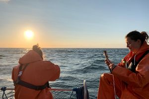 Zwei Frauen in orangen Regenmänteln und -hosen sitzen auf einem Boot und benutzen verschiedene Messgeräte. Im Hintergrund geht die Sonne über dem Meer unter.