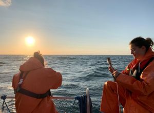 Zwei Frauen in orangen Regenmänteln und -hosen sitzen auf einem Boot und benutzen verschiedene Messgeräte. Im Hintergrund geht die Sonne über dem Meer unter.