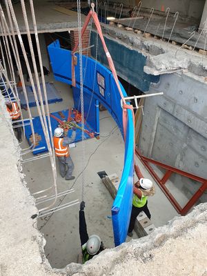 Blick auf eine gebogene Scheibe, eingepackt in blaue Folie, und mehrere Baustellenarbeitskräfte in Warnwesten mit Helmen im Untergeschoss durch eine Deckenöffnung im Erdgeschoss auf einer Baustelle.