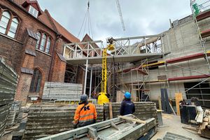 Blick auf eine Baustelle, zwei Personen mit Bauhelm schauen nach oben in Richtung eines Krans