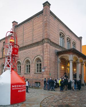 Eine Gruppe von Menschen steht an einer Boje, auf der der Schriftzug "Meeresmuseum" steht