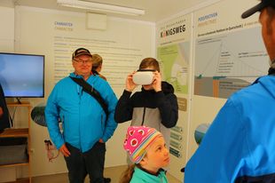 In der Ausstellung im Nationalparkzentrum stehen mehrere Mitglieder. Im Hintergrund sind Wandtafeln mit Informationen zum Königsweg. In der Mitte steht ein Jugendlicher mit einer VR-Brille.
