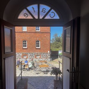 Eine offene Tür gibt den Blick auf einen sonnigen Innenhof mit Kopfsteinpflaster und Backsteinbau frei