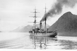 Historisches Bild in schwarz-weiß zeigt ein Schiff mit rauchendem Schornstein auf dem Wasser. Im Hintergrund ist eine Bergkette zu sehen.