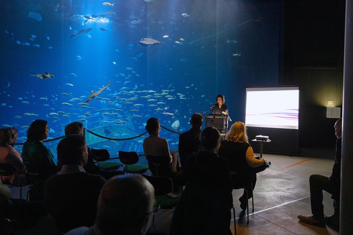 Ein Publikum in einer Vortragssituation vor einer bodentiefen Aquarienscheibe