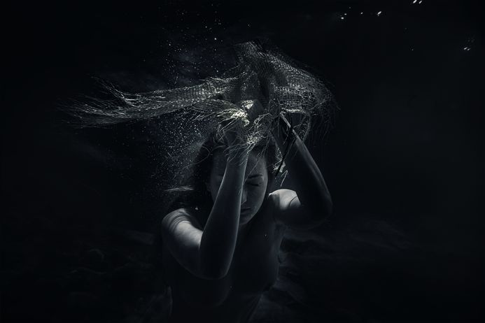Ein dunkles Bild mit einer Frau unter Wasser mit einem Netz in den Händen über dem Kopf. 