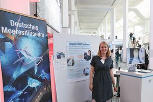 Eine Frau an einem Stand mit Aufstellern, Plakaten und Bildschirmpräsentation.