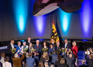 Personen mit Blumensträußen stehen vor einer Flagge mit dem Deutschen Bundesadler