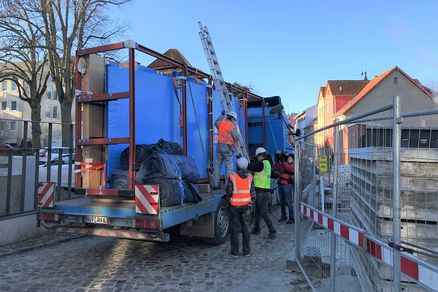 Blick auf einem LKW mit in blauer Folie eingepackten Aquarienscheiben auf der Ladefläche. Anbei stehen Arbeitskräfte in Baustellenbekleidung mit einer Leiter.