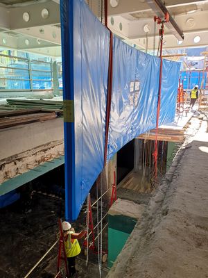 Eine in blaue Folie eingepackte Acrylglasscheibe für ein Aquarium hängt über einer Deckenöffnung im Erdgeschoss auf einer Baustelle.