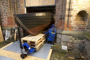Die Vorderseite eines schwarzen Holzbootes auf Palletten und Transportfahrzeug inmitten einer Türöffnung eines Backsteingebäudes.