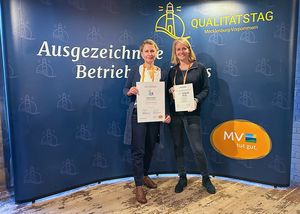 Zwei Frauen stehen lächelnd vor einem blauen Hintergrund und halten Urkunden in den Händen