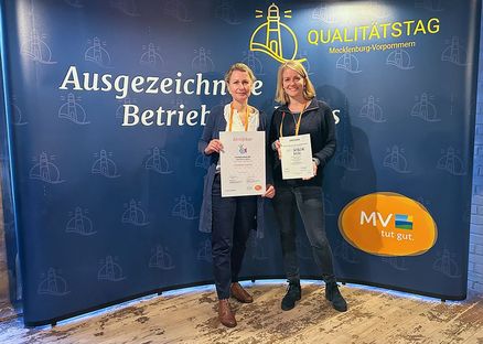 Zwei Frauen stehen lächelnd vor einem blauen Hintergrund und halten Urkunden in den Händen