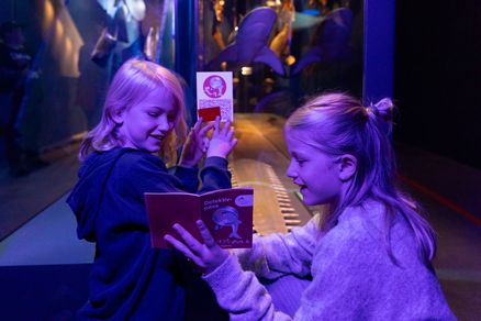 Zwei Kinder stehen unter UV-Licht vor einer Vitrine und halten kleine Hefte in den Händen. Sie zeigen sich gegenseitig die Hefte und erkunden es gemeinsam.