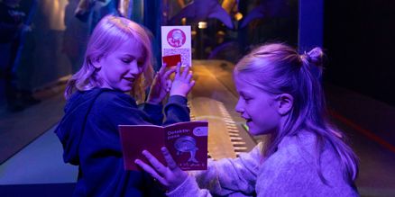 Zwei Kinder stehen unter UV-Licht vor einer Vitrine und halten kleine Hefte in den Händen. Sie zeigen sich gegenseitig die Hefte und erkunden es gemeinsam.