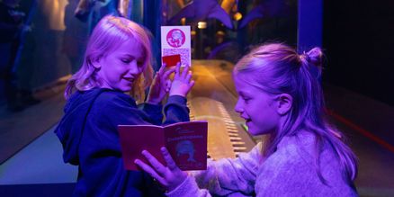 Zwei Kinder stehen unter UV-Licht vor einer Vitrine und halten kleine Hefte in den Händen. Sie zeigen sich gegenseitig die Hefte und erkunden es gemeinsam.
