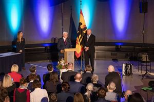 Zwei Personen stehen vor einer Flagge mit dem Deutschen Bundesadler in einem blaubleuchtetem Raum vor Publium