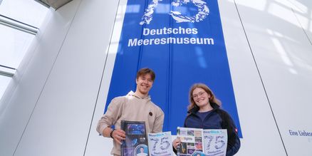 Zwei Personen halten Zeitungen in den Händen und stehen vor einem blauen Banner
