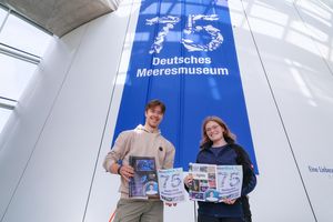 Zwei Personen halten Zeitungen in den Händen und stehen vor einem blauen Banner