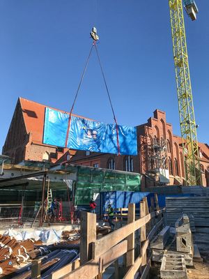 Ein Turmdrehkran hebt eine Scheibe über die Baustelle. Im Hintergrund ist ein altes Backsteingebäude sichtbar. 