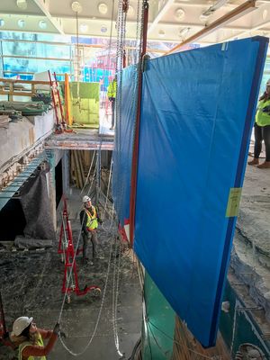 Eine in blaue Folie eingepackte, gebogene Aquarienscheibe wird durch eine Deckenöffnung im Erdgeschoss ins Untergeschoss einer Baustelle herabgelassen. 