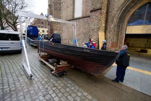 Einige Männer stehen um ein schwarzes Holzboot, das mit einer Hebekonstruktion vor einem Backsteingebäude in Richtung eines LKWs transportiert wird.
