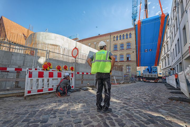 Zwei Männer schauen zu, wie eine riesige Acrylglasscheibe vorsichtig von zwei Kränen in der Luft aufgerichtet wird. (Foto: Anke Neumeister/Deutsches Meeresmuseum)