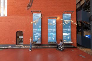 A standing woman and a man sitting in a wheelchair are inside a historic brick building.