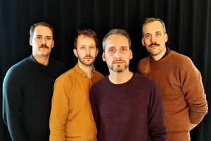 Vier Männer in farbigen Pullovern stehen vor einer schwarzen Wand.