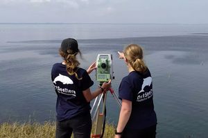 Zwei Frauen haben den Rücken zur Kamera gedreht und stehen am Ufer eines großen Gewässers. Sie blicken zum Horizont, zwischen ihnen steht ein Vermessungsgerät.