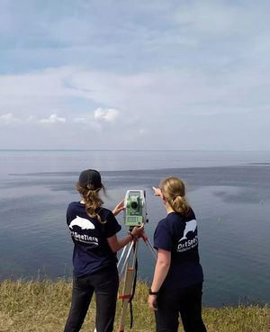 Zwei Frauen haben den Rücken zur Kamera gedreht und stehen am Ufer eines großen Gewässers. Sie blicken zum Horizont, zwischen ihnen steht ein Vermessungsgerät.