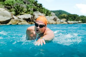 Eine Person mit Taucherbrille und oranger Badekappe schwimmt in türkisblauem Meer. Im Hintergrund ist eine felsige Küstenlandschaft zu sehen.