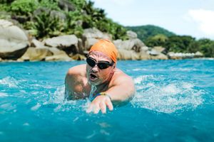 Eine Person mit Taucherbrille und oranger Badekappe schwimmt in türkisblauem Meer. Im Hintergrund ist eine felsige Küstenlandschaft zu sehen.