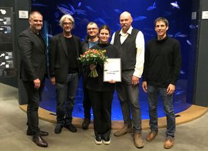 Sechs Menschen stehen vor einem Aquarium, in der Mitte ist die Preisträgerin der "Forschungsstiftung Ostsee" mit Blumenstrauß und Urkunde.