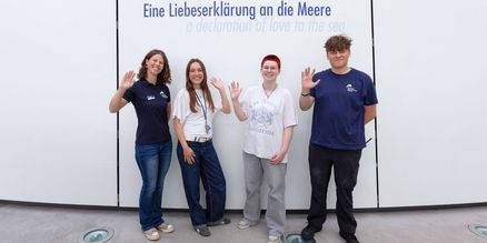 Vier junge Erwachsene stehen lächelnd vor einer weißen Wand auf der die Aufschrift "Eine Liebeserklärung an die Meere" zu lesen ist. 