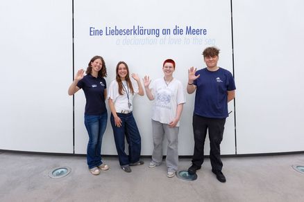 Vier junge Erwachsene stehen lächelnd vor einer weißen Wand auf der die Aufschrift "Eine Liebeserklärung an die Meere" zu lesen ist. 