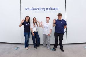 Vier junge Erwachsene stehen lächelnd vor einer weißen Wand auf der die Aufschrift "Eine Liebeserklärung an die Meere" zu lesen ist. 