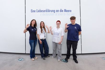 Vier junge Erwachsene stehen lächelnd vor einer weißen Wand auf der die Aufschrift "Eine Liebeserklärung an die Meere" zu lesen ist. 